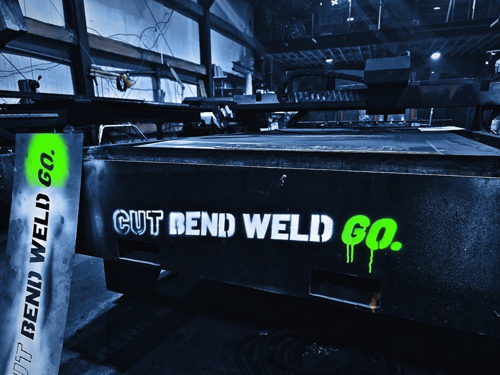 The Cut Bend Weld Go logo sprayed in white and lime on the side of the plasma table, the cut steel stencil leaning beside it