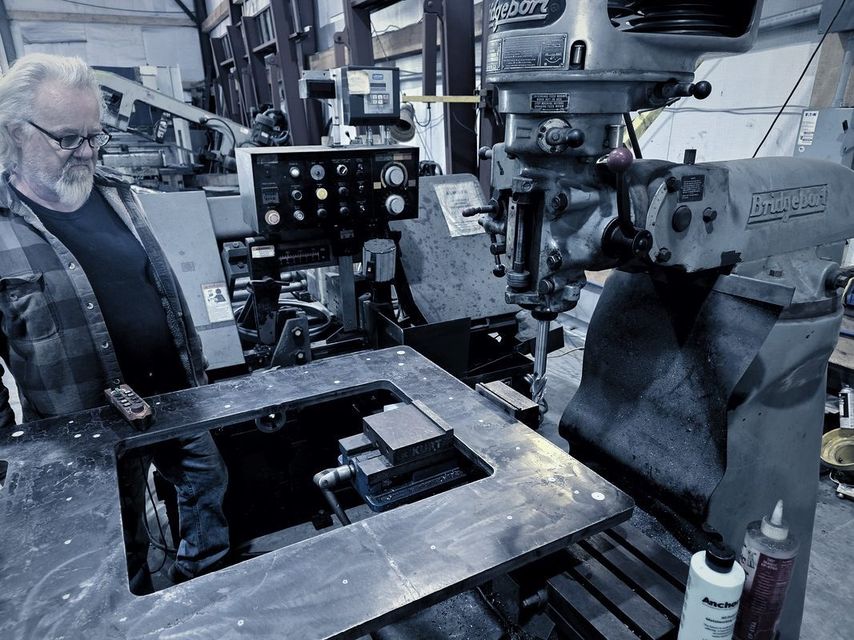 A man with a white beard stands at the milling machine over a plate with a square opening