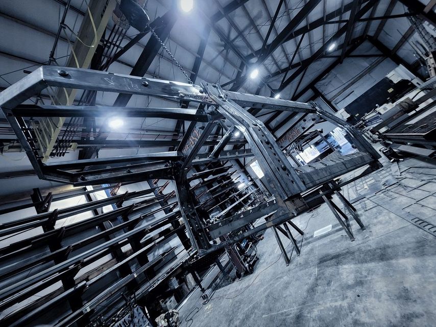 A large welded steel frame hangs from a chain hoist in the shop