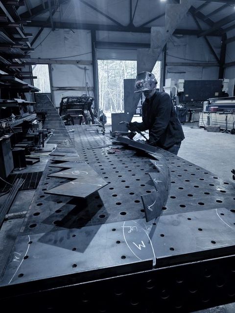 A welder in a hood works on curved stair parts laid out on the fixture table