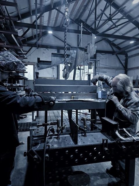 Two men fit a steel beam on the fixture table while a stair section hangs from the hoist
